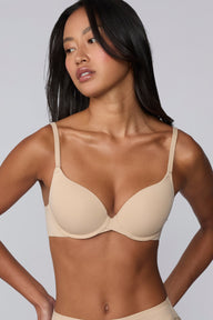 Against a light grey background, Amber models the Base Push-Up Bra in Beige, hands at her hips. The molded, lightly lined cups enhance lift and shape, with thin adjustable straps and smooth microfiber fabric for everyday comfort.