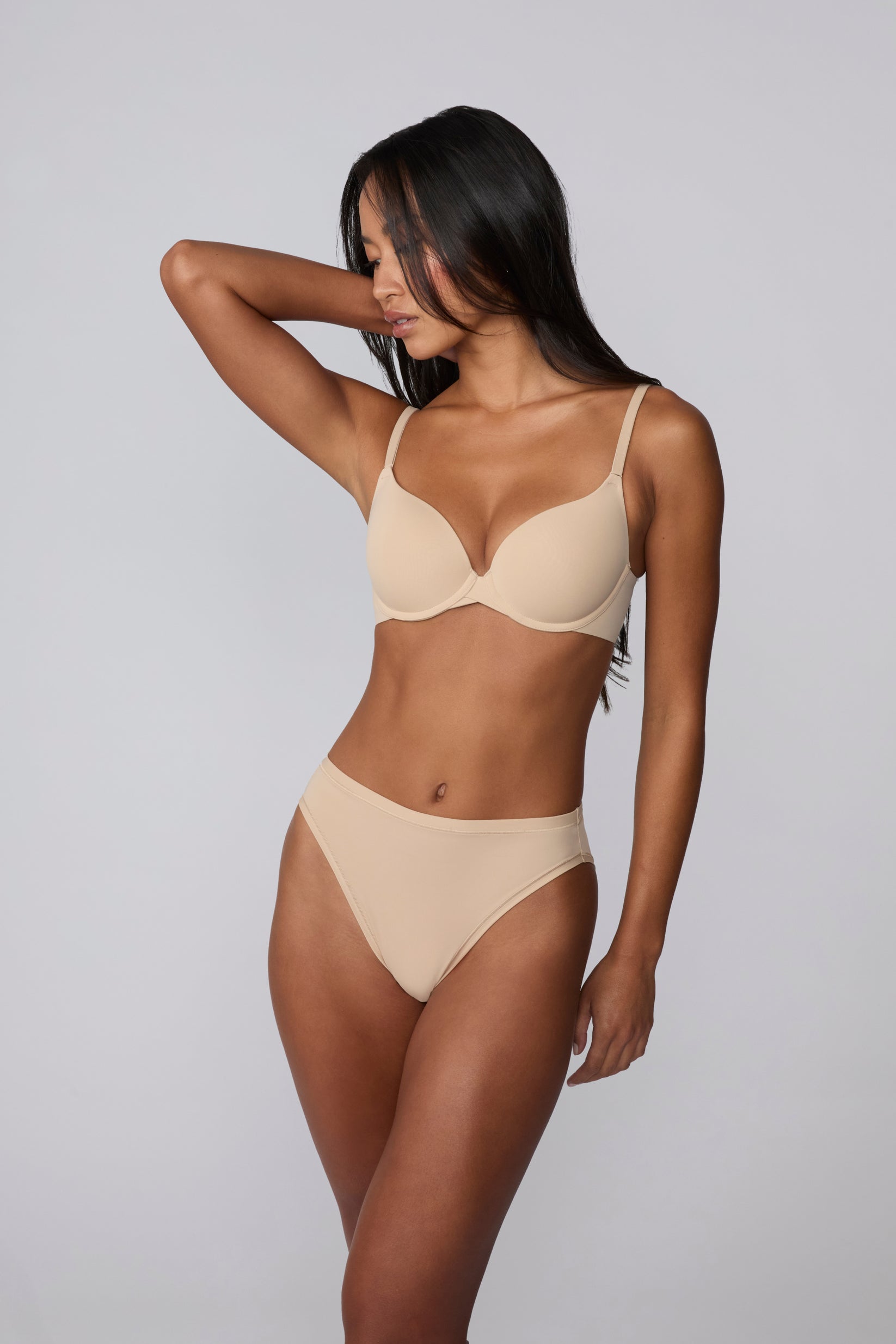 On a light grey studio background, Amber wears the Base Push-Up Bra in Beige with one arm raised. The molded, lightly lined push-up cups create lift and definition, finished with thin adjustable straps and a smooth microfiber band.