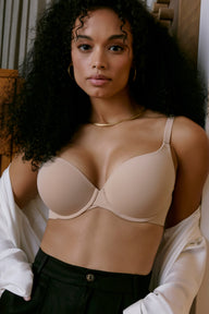 Against a warm-toned interior backdrop, a model wears the Base Push-Up Bra in Beige with black trousers and a draped white shirt. The lightly lined underwire cups create lift and shape, finished with a smooth microfiber band and adjustable straps. sizing: Bree wears size 32E