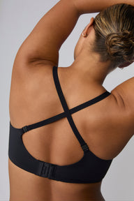 On a light grey studio backdrop, India wears the Base Push-Up Bra in Black from the back with straps crossed. The convertible straps form an X shape, and the microfiber band features a hook-and-eye closure.