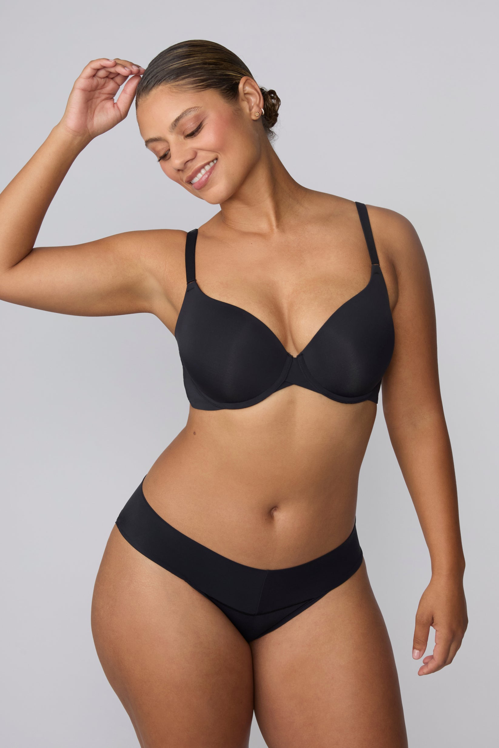 On a light grey studio backdrop, India wears the Base Push-Up Bra in Black with matching underwear. The lightly lined underwire cups create lift and shape, finished with thin adjustable straps and a smooth microfiber band.