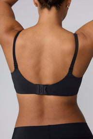 Against a light grey background, India models the Base Push-Up Bra in Black from the back. The adjustable straps and hook-and-eye closure are visible, with the smooth microfiber band laying flat across the back.