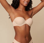 Three quarter view of Ally posing with her hands behind her head, modeling the Base Strapless Bra in Beige against a simple, neutral background.