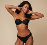 Full-body view of Ally posing with her hands behind her head, modeling the Base Strapless Bra in Black against a simple, neutral background.