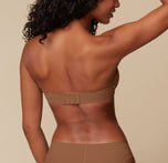 Back view of Ally modeling the Base Strapless Bra in Brown. She poses with her arms lifted, emphasizing the back closure and smooth band of the strapless bra.