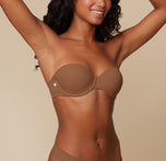 Three quarter view of Ally posing with her hands behind her head, modeling the Base Strapless Bra in Brown against a simple, neutral background.
