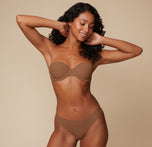 Full-body view of Ally posing with her hands behind her head, modeling the Base Strapless Bra in Brown against a simple, neutral background.