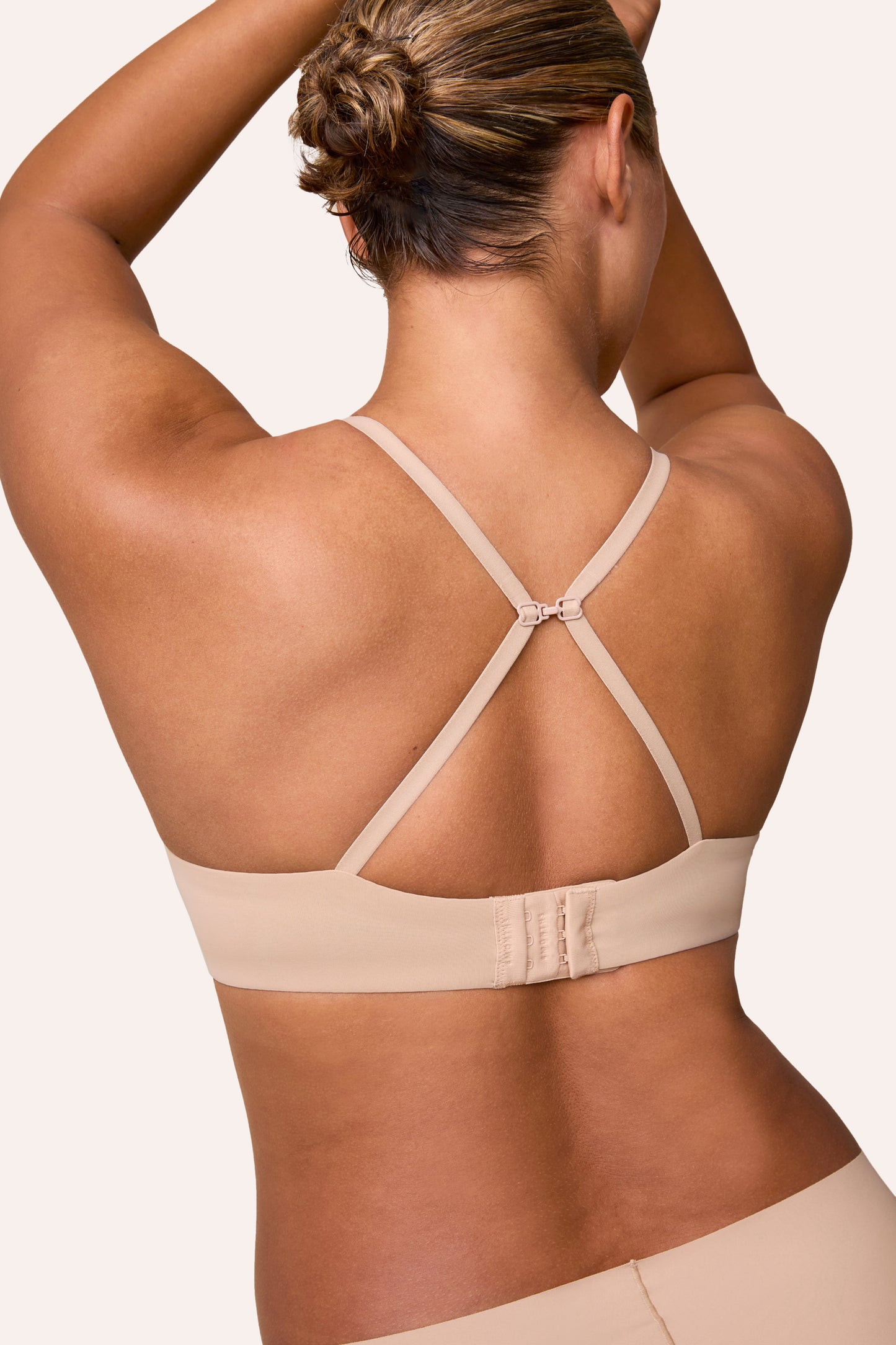 On a light neutral studio backdrop, India wears the Base T-Shirt Bra in Beige, shown from the back in a racerback position. The J-hook connects the straps for a convertible fit, and the microfiber band sits smooth across the back with a hook-and-eye closure.