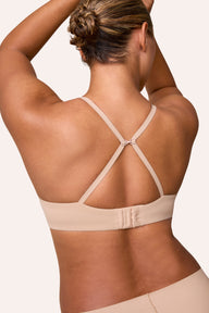 On a light neutral studio backdrop, India wears the Base T-Shirt Bra in Beige, shown from the back in a racerback position. The J-hook connects the straps for a convertible fit, and the microfiber band sits smooth across the back with a hook-and-eye closure.