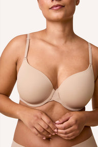 On a light neutral studio backdrop, India wears the Base T-Shirt Bra in Beige, shown from the front. The lightly lined cups create smooth, full coverage, and the front-adjusting straps and signature X-bridge are visible at the center.