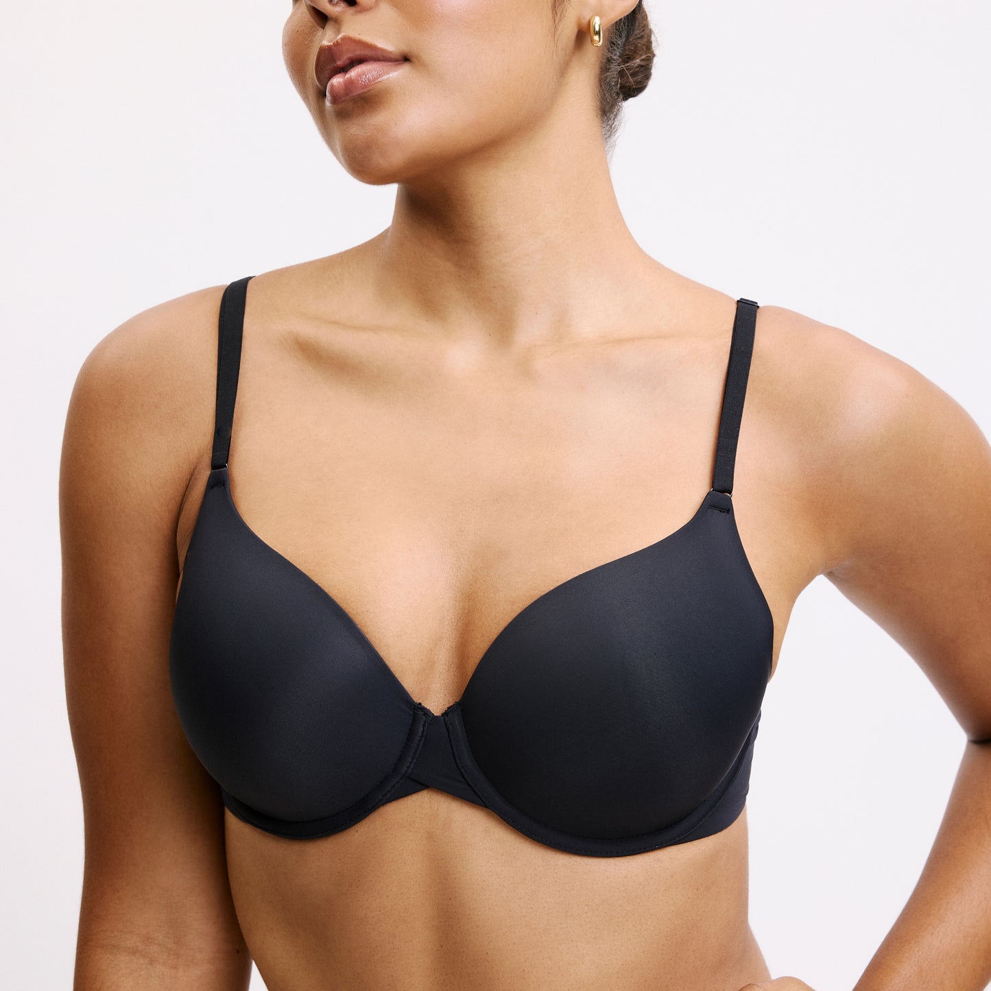 On a light studio backdrop, Amber wears the Base T-Shirt Bra in Black in a cropped close-up view. The lightly lined underwire cups and smooth microfiber fabric create clean coverage under thin front-adjusting straps.