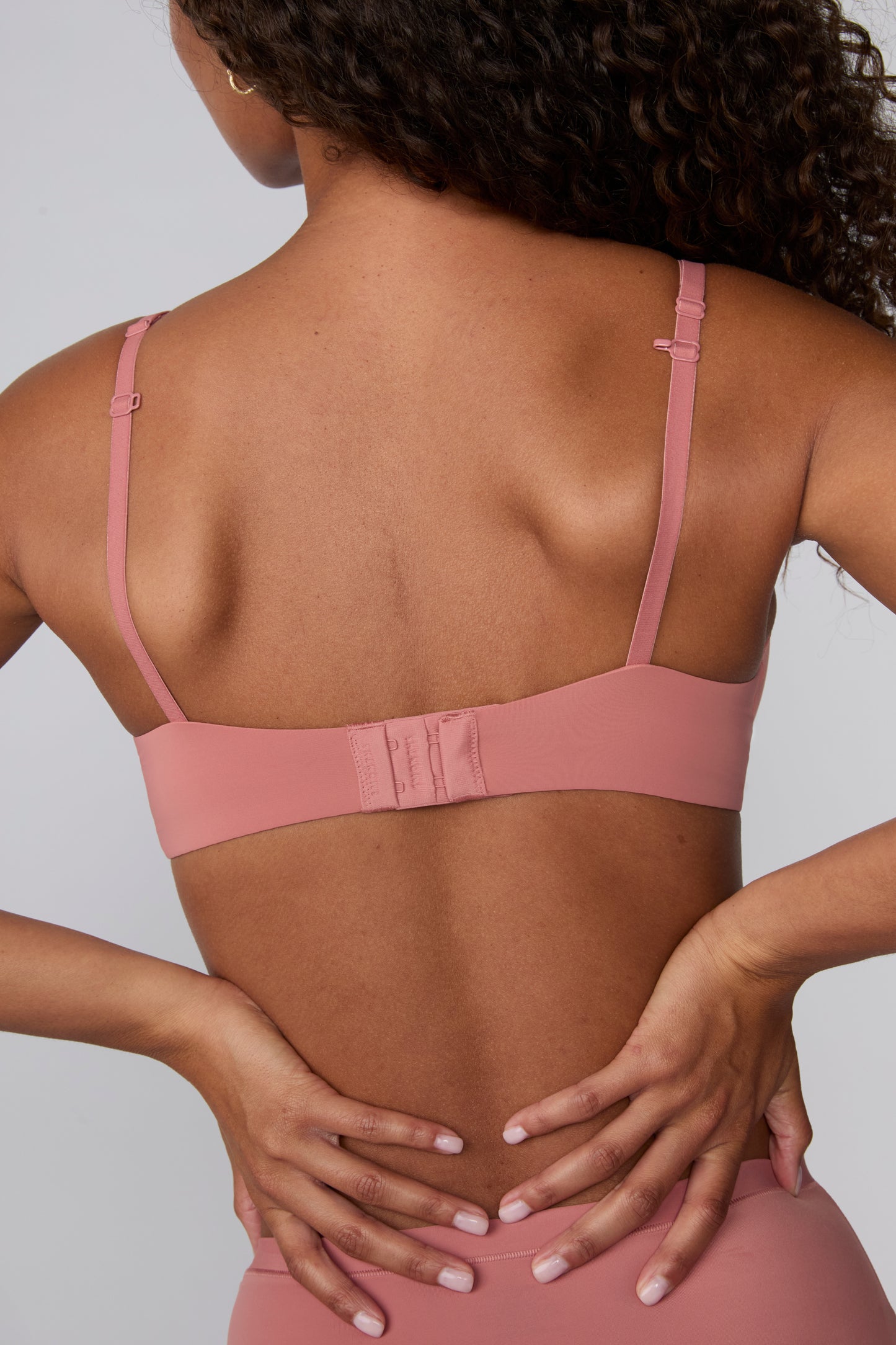 In a studio with a light gray backdrop, Courtney wears the Base T-Shirt Bra in Cinnamon from the back, highlighting the smooth microfiber band, adjustable straps, and hook-and-eye closure.