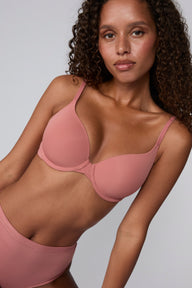 In a studio with a light gray backdrop, Courtney wears the Base T-Shirt Bra in Cinnamon, showing the lightly lined underwire cups, smooth microfiber fabric, and front-adjusting straps.