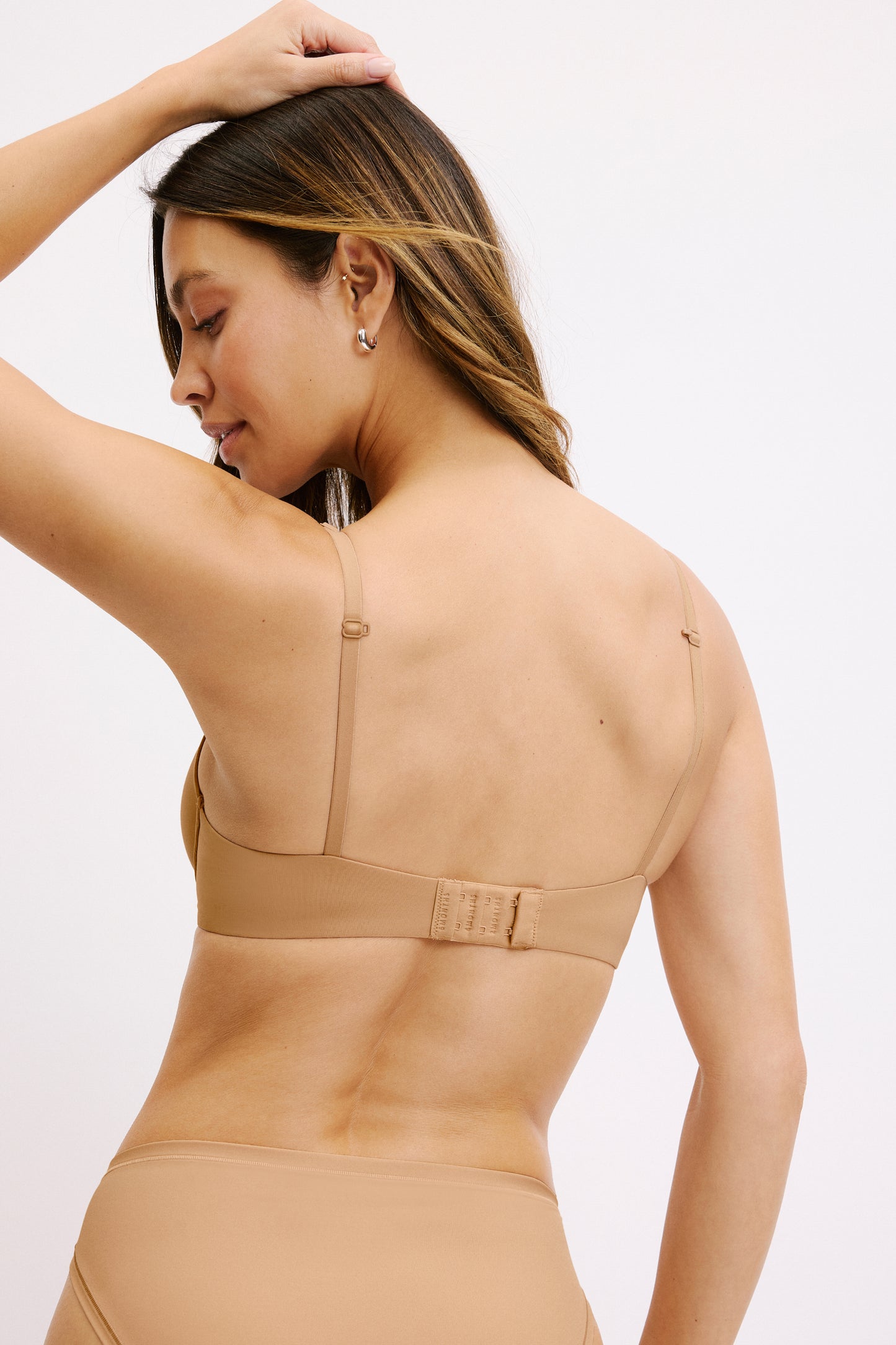 On a white studio backdrop, Darion wears the Base T-Shirt Bra in Tan from the back, showing the adjustable straps, smooth microfiber band, and hook-and-eye closure.