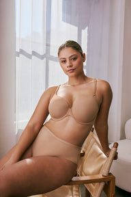 In bright natural light near a window, a model sits on a chair wearing the Base T-Shirt Bra in Tan paired with matching high-waist underwear, showing lightly lined cups, adjustable straps, and smooth microfiber fabric.