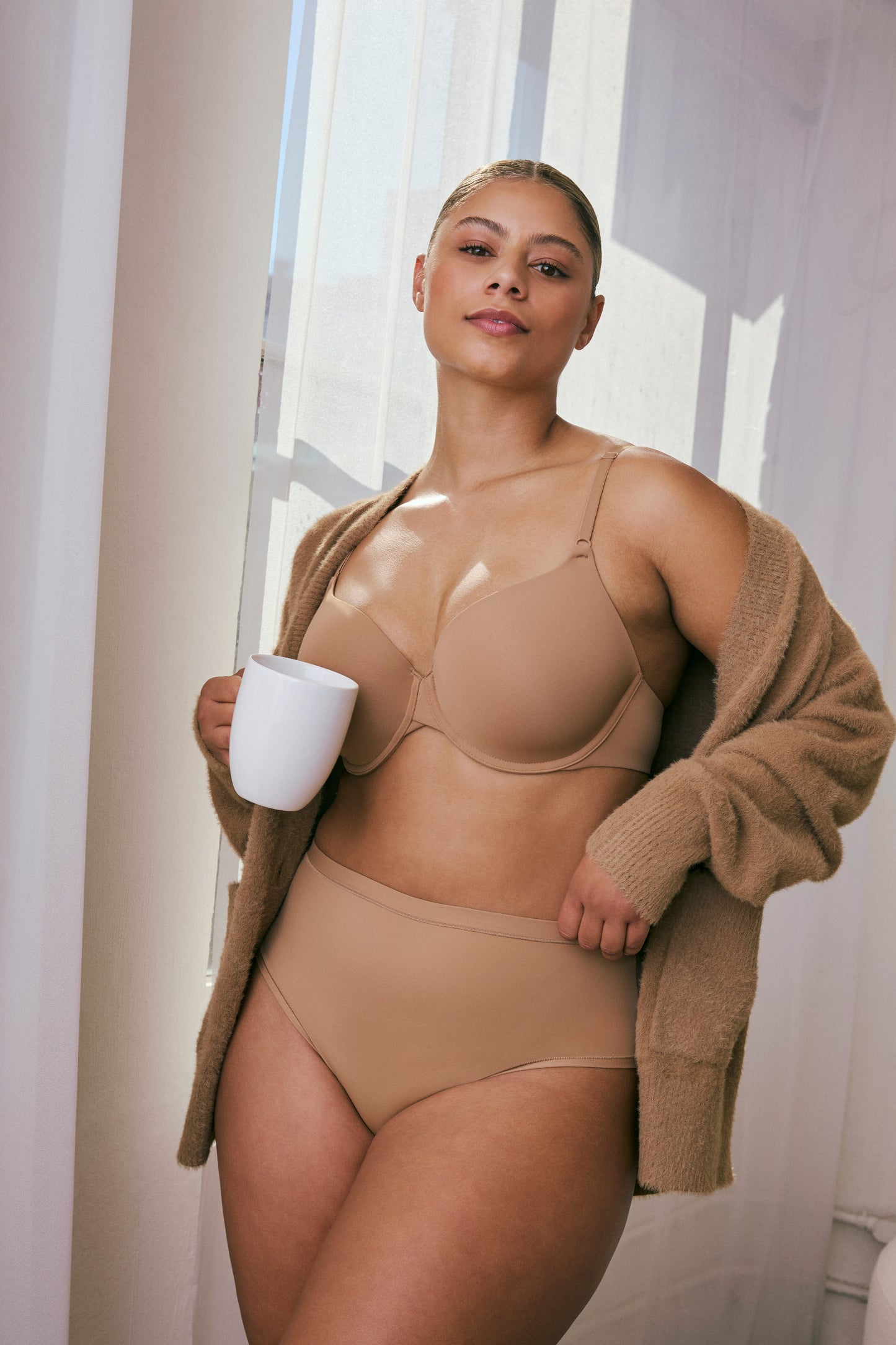 In bright natural light near a window, a model wears the Base T-Shirt Bra in Tan paired with matching high-waist underwear, showing lightly lined cups, adjustable straps, and smooth microfiber fabric. She holds a white mug and wears a soft cardigan draped over her shoulders.