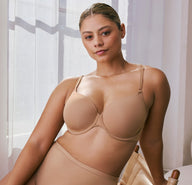 In a softly sunlit room with sheer curtains, a model wears the Base T-Shirt Bra in Tan, highlighting the lightly lined underwire cups, smooth microfiber fabric, and front-adjusting straps. She is seated in a wooden chair, styled with matching high-waist underwear.