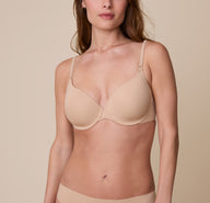 Gracie wears the Base T-Shirt Bra in Beige, facing forward with a soft expression. The image highlights the smooth, lightly lined cups, underwire support, and adjustable straps against a neutral beige background. sizing: Gracie wears size 32D