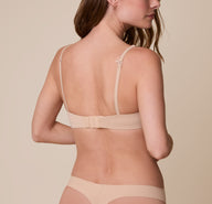 Gracie showcases the back of the Base T-Shirt Bra in Beige, revealing the wide band, hook-and-eye closure, adjustable straps, and convertible racerback feature for a secure fit.