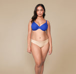 A front view of Claudia standing confidently while wearing the Base Unlined Bra in Deja Blue and the No-Show Bikini in Beige. The image highlights the full set, showcasing the bold color of the bra and the neutral tone of the bikini.