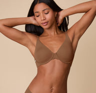Ally poses with her arms behind her head, wearing the Base Unlined Bra in Brown. The image highlights the bra’s deep V neckline, smooth microfiber fabric, and lightly supportive underwire design. sizing: Ally wears size 32D