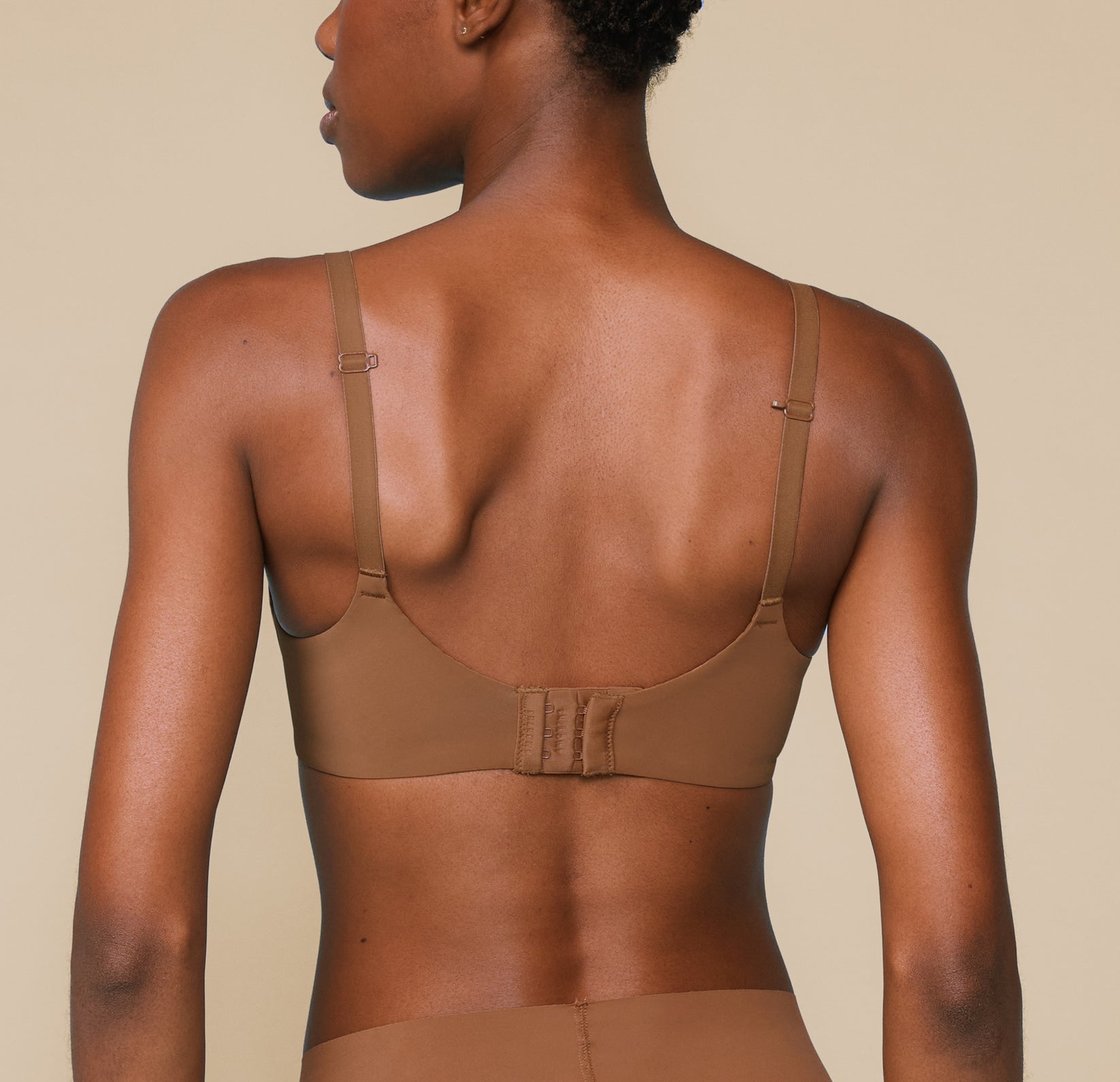 Back view of Mariah wearing the Bliss Unlined Bra in Brown. TheJ-Hook convertible racerback feature and hook and eye clasps are highlighted.