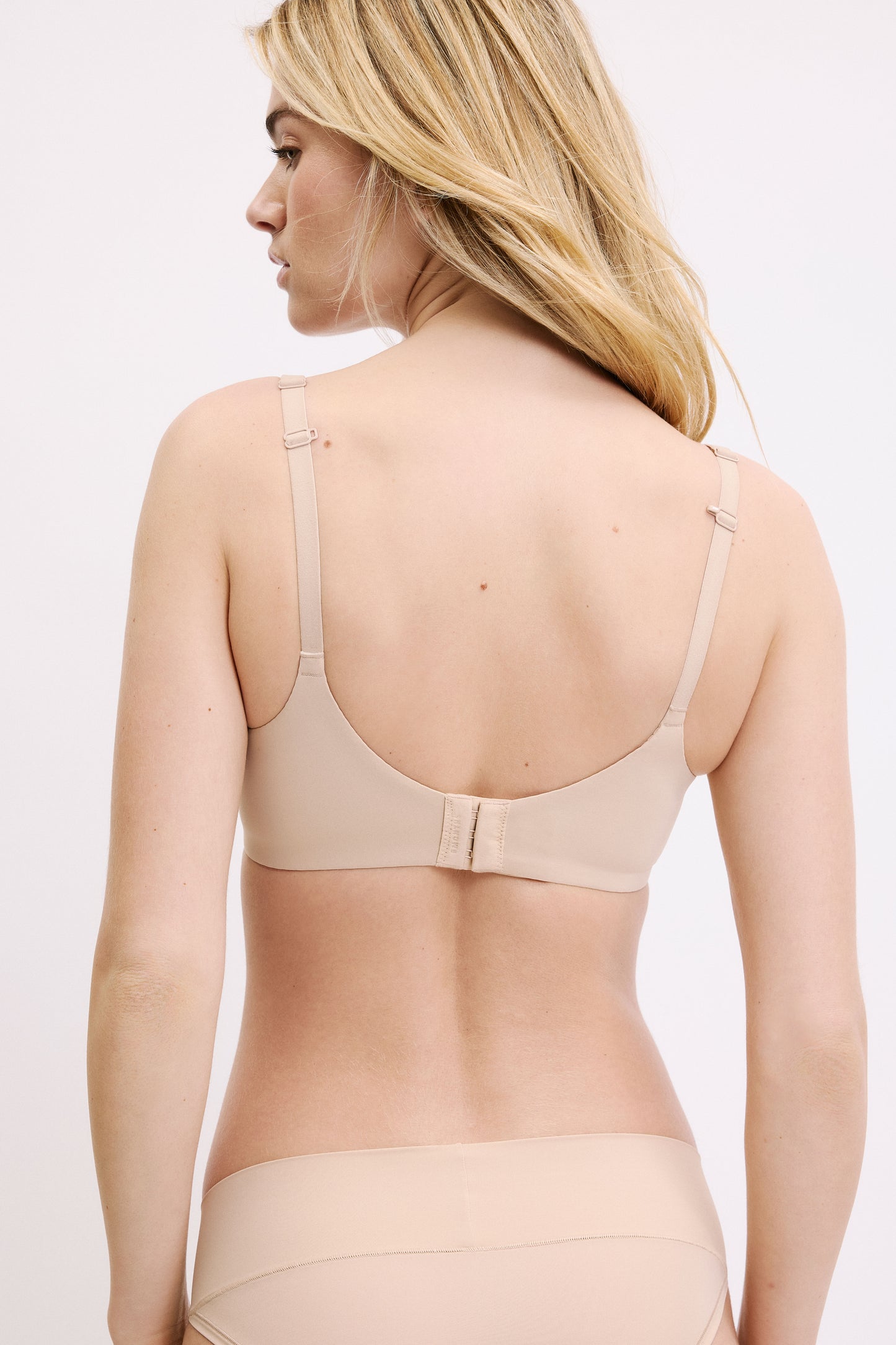 On a white studio backdrop, Megan is shown from behind wearing the Base Unlined Bra in Beige, highlighting the adjustable straps configured into a racerback and the hook-and-eye closure.