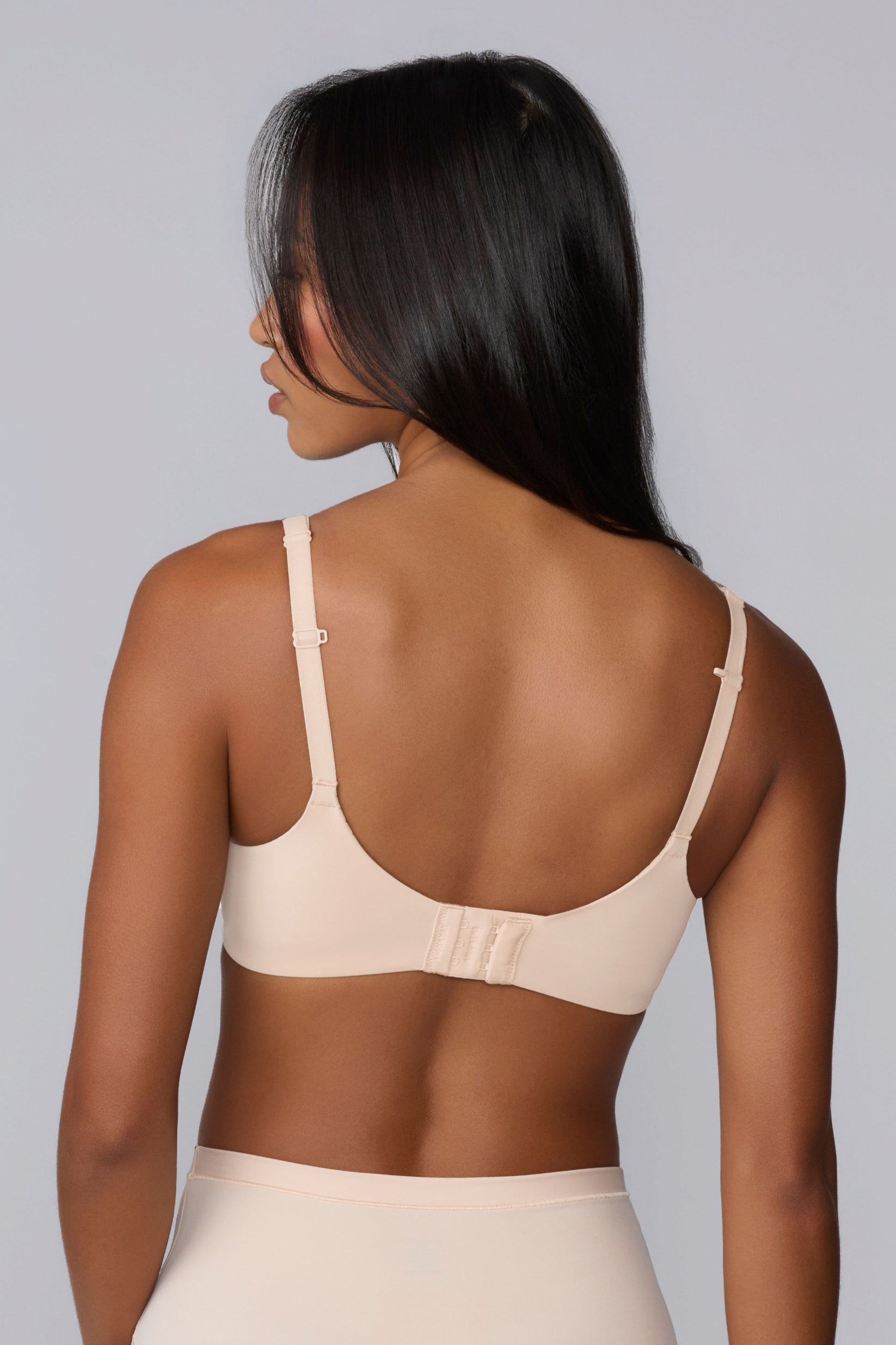 In a studio with a light gray backdrop, Amber wears the Base Unlined Bra in Rose from the back, highlighting the smooth microfiber band, adjustable straps, and hook-and-eye closure.