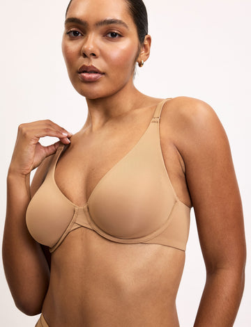 On a light studio backdrop, Amber wears the Base Unlined Bra in Tan, featuring unlined underwire cups, adjustable straps, and a smooth microfiber finish. sizing: Amber wears size 34D