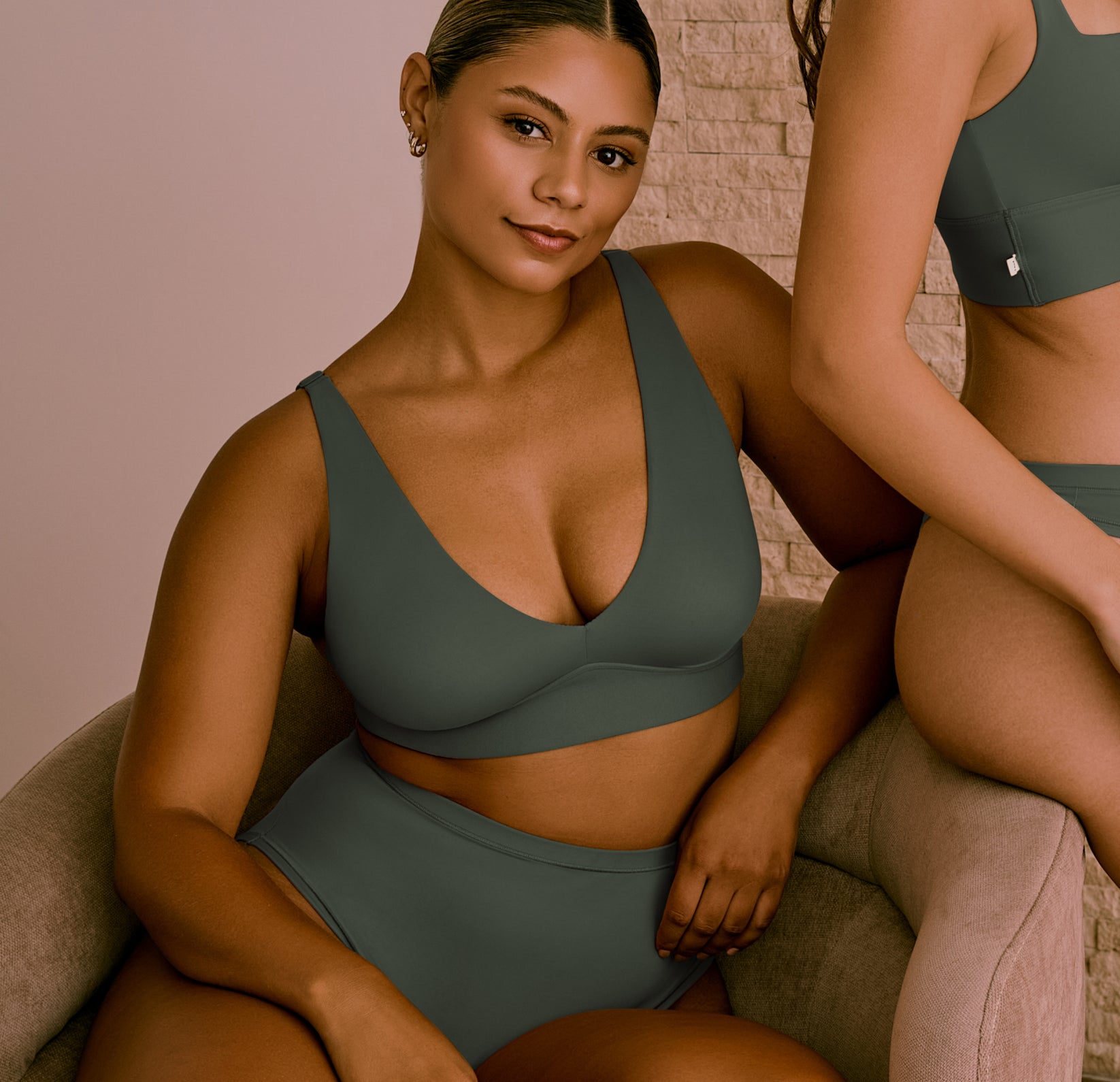 Two models wear Bliss Collection pieces in Fir. The seated model wears the Bliss Adjustable Bralette and Bliss Highwaist Brief, showcasing the deep V neckline and high-rise coverage. The standing model wears the Bliss Square Neck Bralette and matching brief.