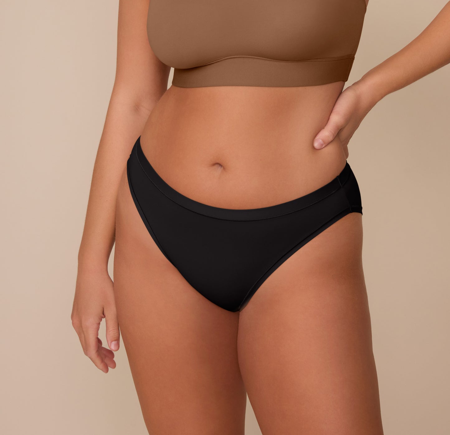 Close-up front view of Sonnie wearing the Bliss Bikini in Black and the Bliss Scoop Bralette in Brown. The bikini’s high-rise, mid-cut design offers moderate coverage and a smooth fit against the skin. sizing: Sonnie wears size Large