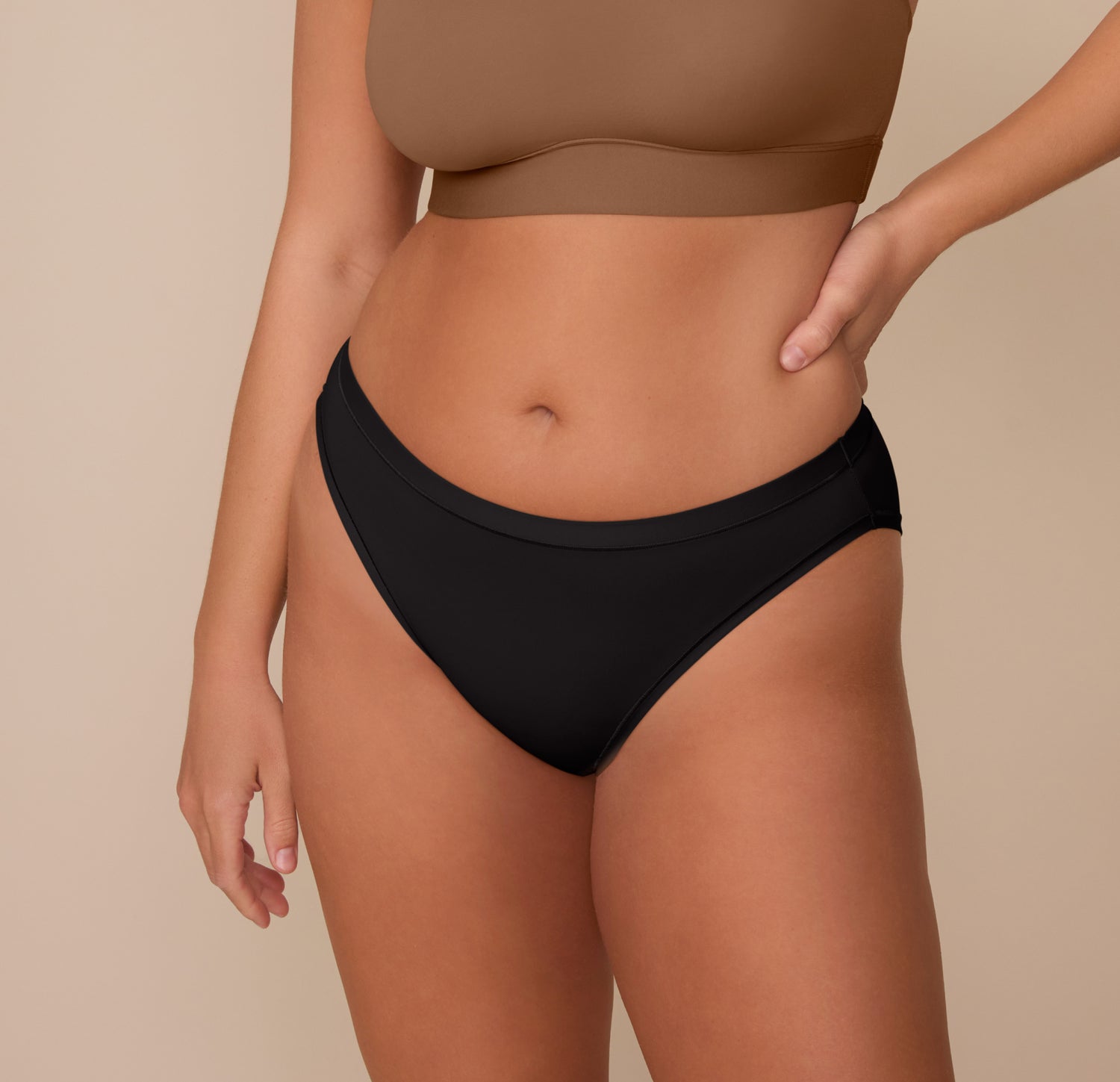 Close-up front view of Sonnie wearing the Bliss Bikini in Black and the Bliss Scoop Bralette in Brown. The bikini’s high-rise, mid-cut design offers moderate coverage and a smooth fit against the skin. sizing: Sonnie wears size Large