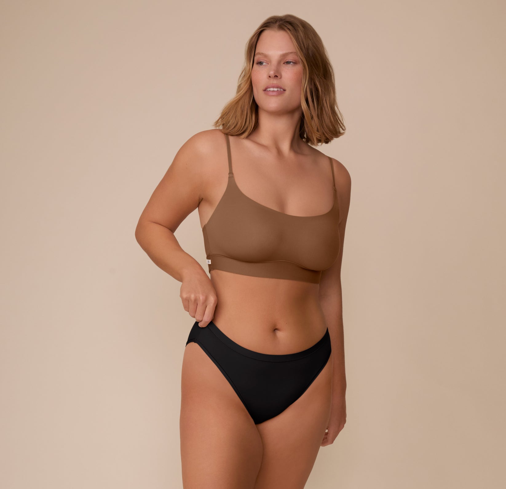 Sonnie stands relaxed, smiling slightly while adjusting the waistband of the Bliss Bikini in Black. She pairs it with the Bliss Scoop Bralette in Brown, emphasizing the set’s sleek, minimal aesthetic.
