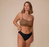 Sonnie stands relaxed, smiling slightly while adjusting the waistband of the Bliss Bikini in Black. She pairs it with the Bliss Scoop Bralette in Brown, emphasizing the set’s sleek, minimal aesthetic.
