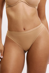 On a light neutral studio backdrop, Amber wears the Bliss Bikini in Tan, shown from the front with the Base T-Shirt Bra. The elastic-free edges and microfiber fabric sit smooth against the skin for no-show comfort. sizing: Amber wears size Large