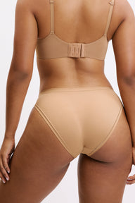 On a light neutral studio backdrop, Amber wears the Bliss Bikini in Tan, shown from the back. The moderate coverage and soft microfiber fabric create a smooth finish under clothing.