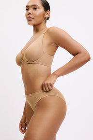 On a light neutral studio backdrop, Amber wears the Bliss Bikini in Tan with the Base T-Shirt Bra, shown in a side angle. The low-rise bikini silhouette and breathable microfiber fabric are designed for everyday comfort.