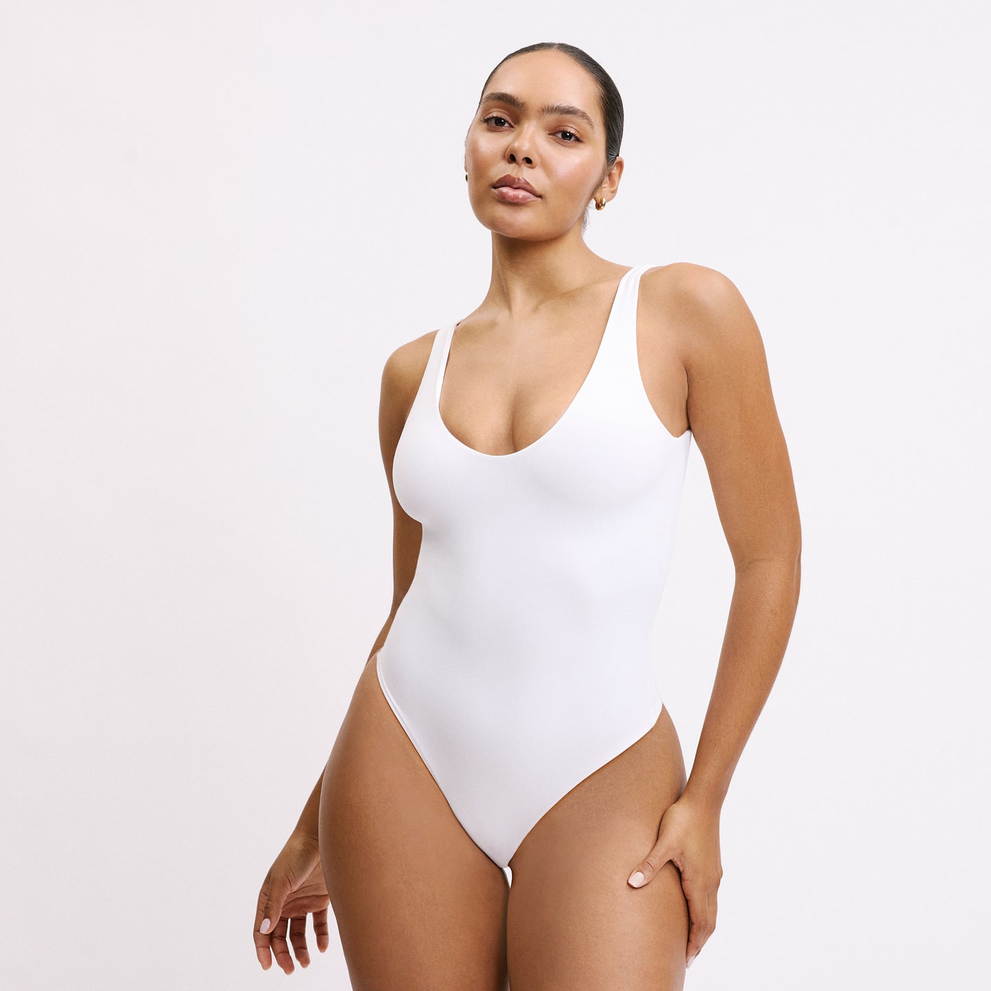 Against a light gray studio backdrop, Amber stands wearing the Bliss Bodysuit in White. The scoop neck, wide straps, and high-cut leg opening showcase the smooth, stretchy microfiber fabric and comfortable pullover design.