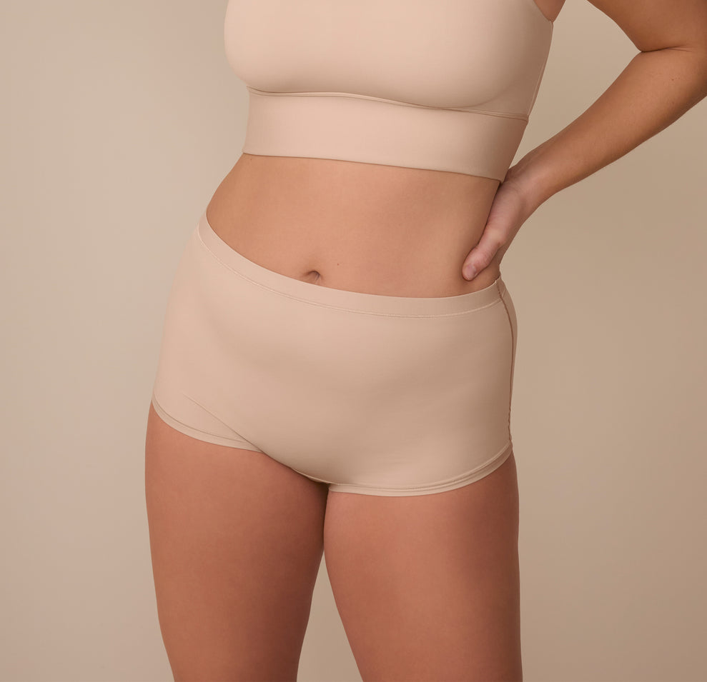 Sonnie wearing the Bliss Boyshort in Beige, standing against a neutral beige background. The boyshort features a mid-rise fit with full coverage and minimal edges for ultimate comfort. sizing: Sonnie wears size XL
