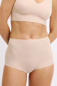 On a white studio backdrop, a close-up shows Darion wearing the Bliss Boyshort in Beige, focusing on the smooth microfiber fabric, high-rise waistband, and clean finish at the leg opening.