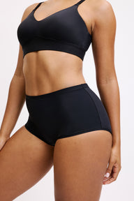On a white studio backdrop, Amber wears the Bliss Boyshort in Black, shown from a slight side angle to highlight the smooth microfiber fabric, mid-rise waistband, and clean edges along the leg opening. sizing: Amber wears size Large