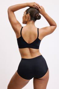 On a white studio backdrop, Amber is shown from behind wearing the Bliss Boyshort in Black with the matching Bliss bralette, highlighting the smooth microfiber fabric, wide waistband, and full back coverage as she lifts her arms overhead.