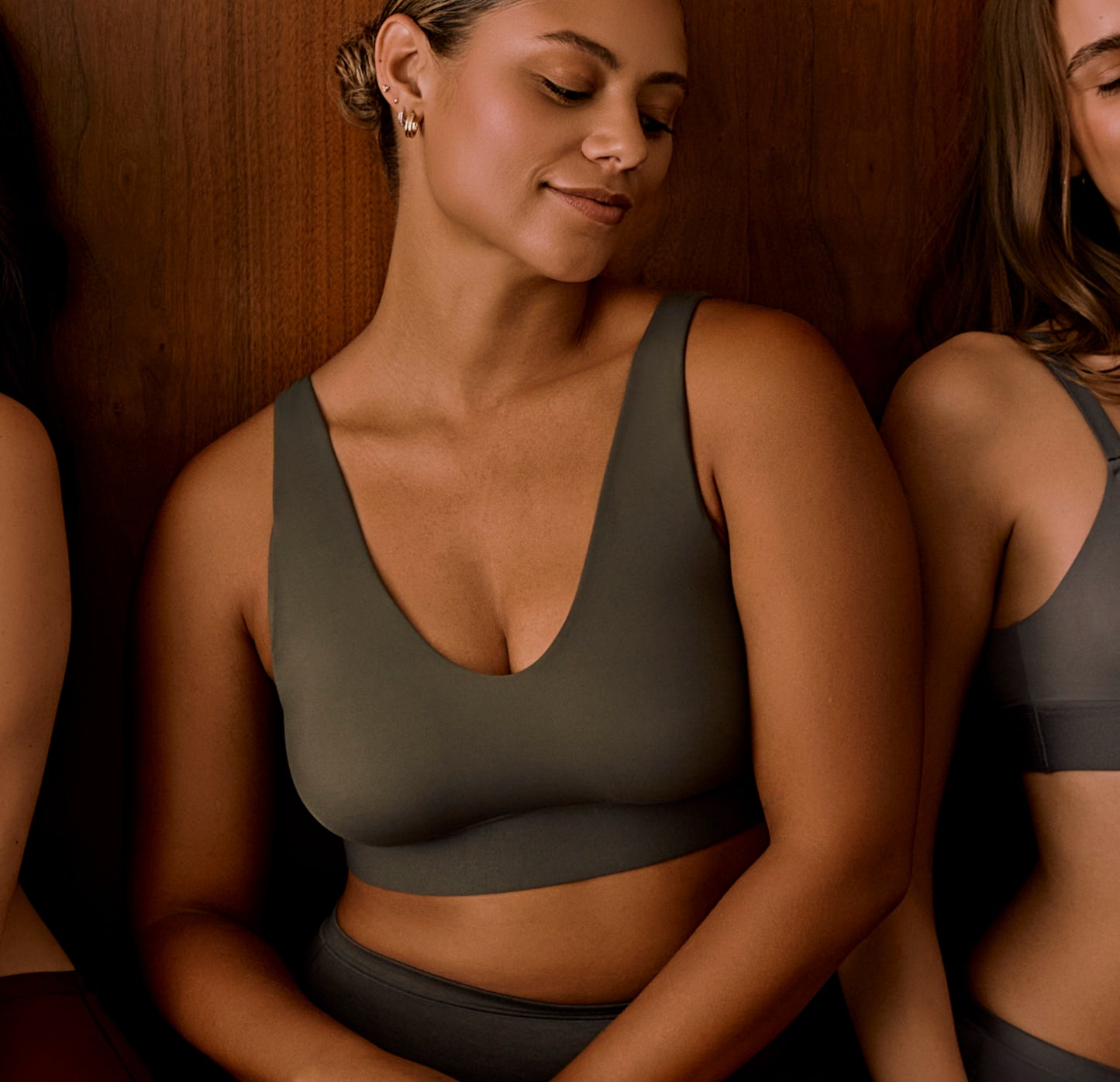 Model wears the Bliss Bralette in Olive, shown from the waist up against a dark wood background. The V neckline and wide straps highlight the smooth, supportive fit.