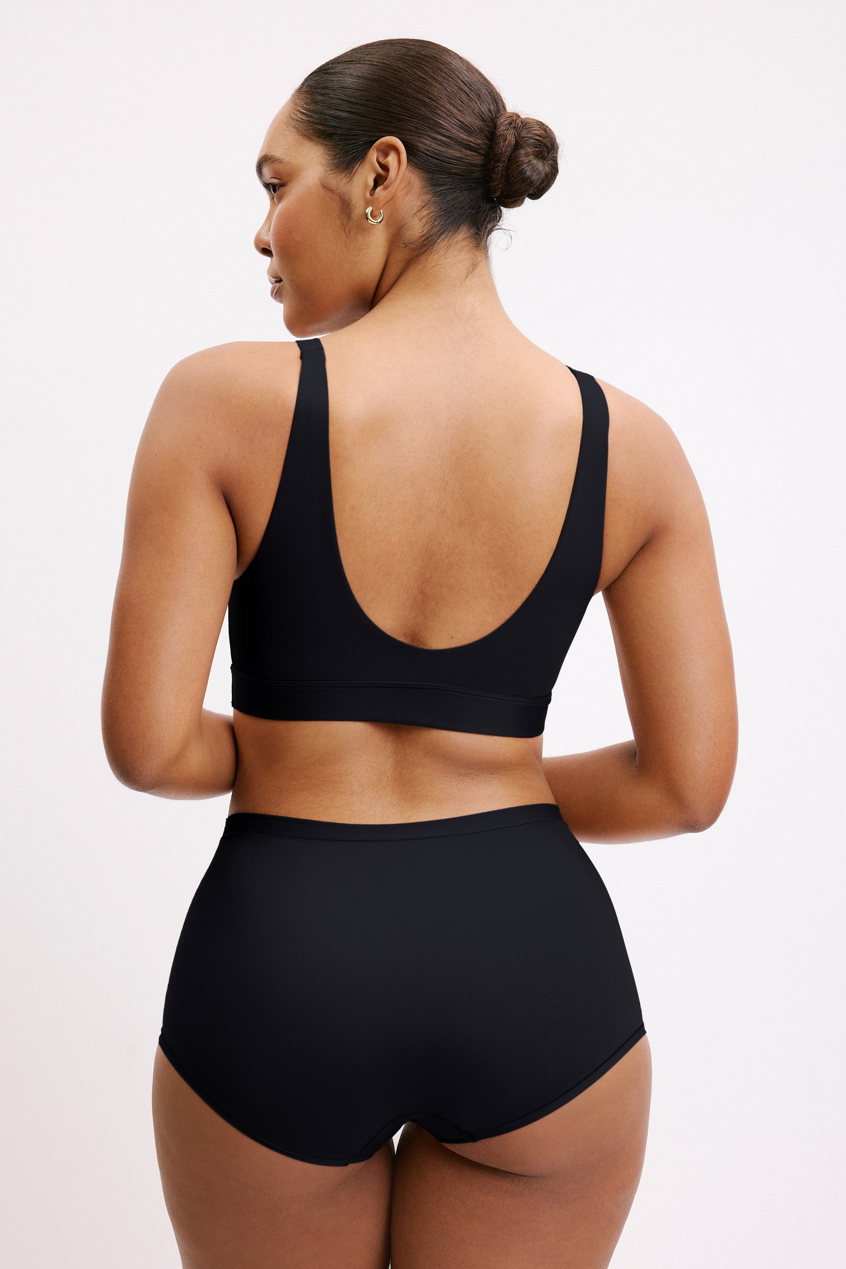 On a light neutral studio backdrop, Amber wears the Bliss Bralette in Black, shown from the back. The wide straps and scoop back provide soft support with a smooth, flexible band.