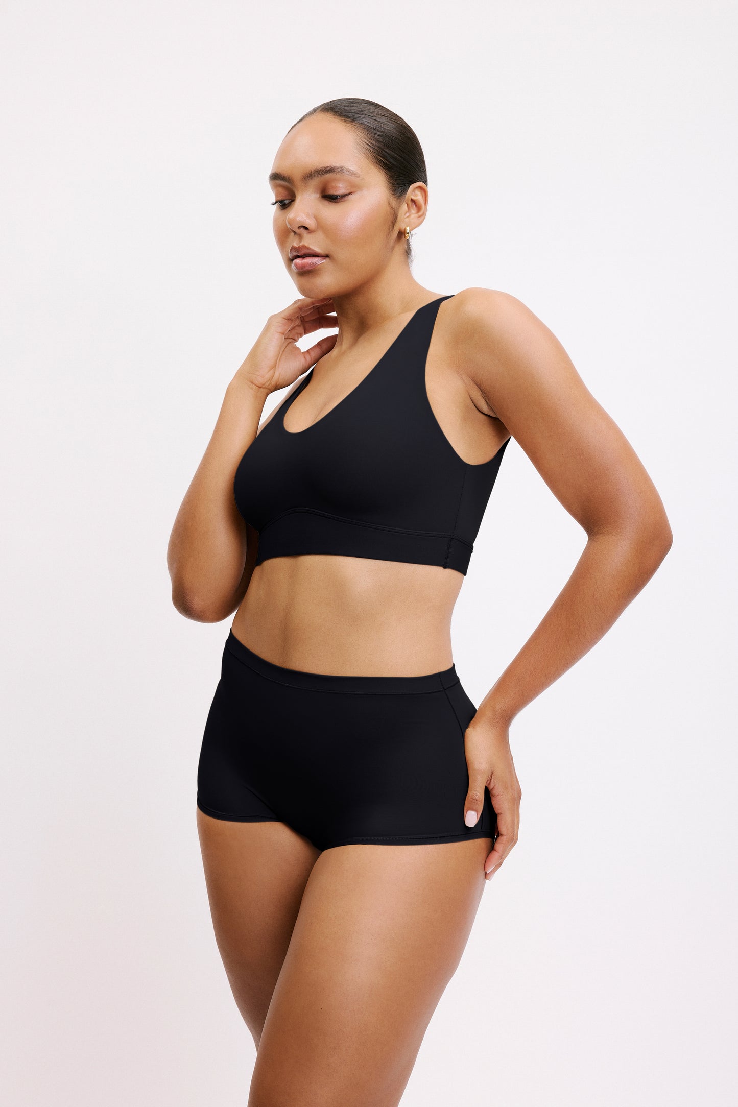 On a light neutral studio backdrop, Amber wears the Bliss Bralette in Black with matching underwear, shown in a three-quarter view. The wireless bralette features a scoop neckline and a supportive underband designed for all-day comfort.