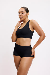 On a light neutral studio backdrop, Amber wears the Bliss Bralette in Black with matching underwear, shown in a three-quarter view. The wireless bralette features a scoop neckline and a supportive underband designed for all-day comfort.