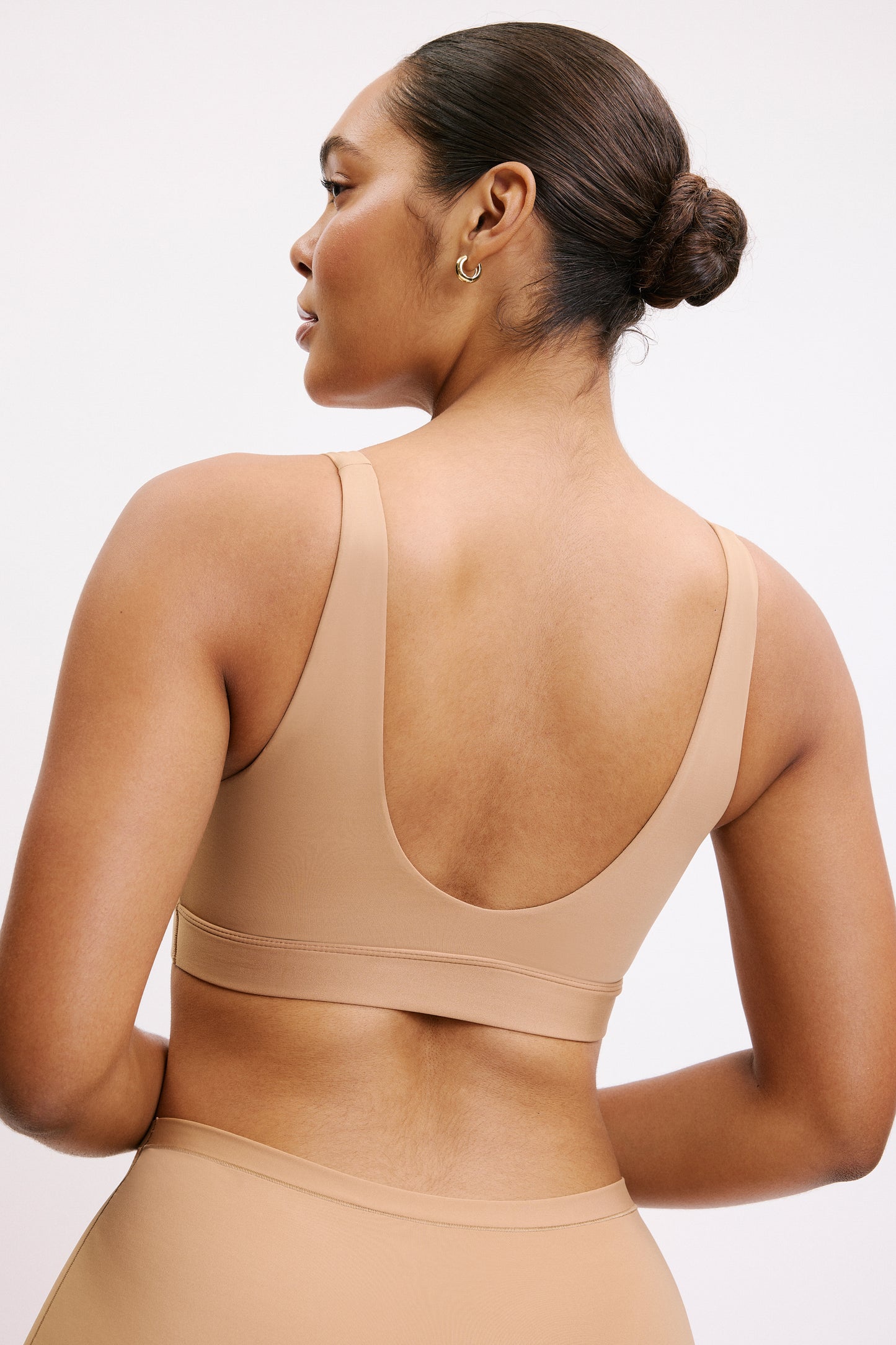 On a white studio backdrop, Amber is shown from the back wearing the Bliss Bralette in Tan, highlighting the wide straps, smooth back band, and clean, wire-free construction.
