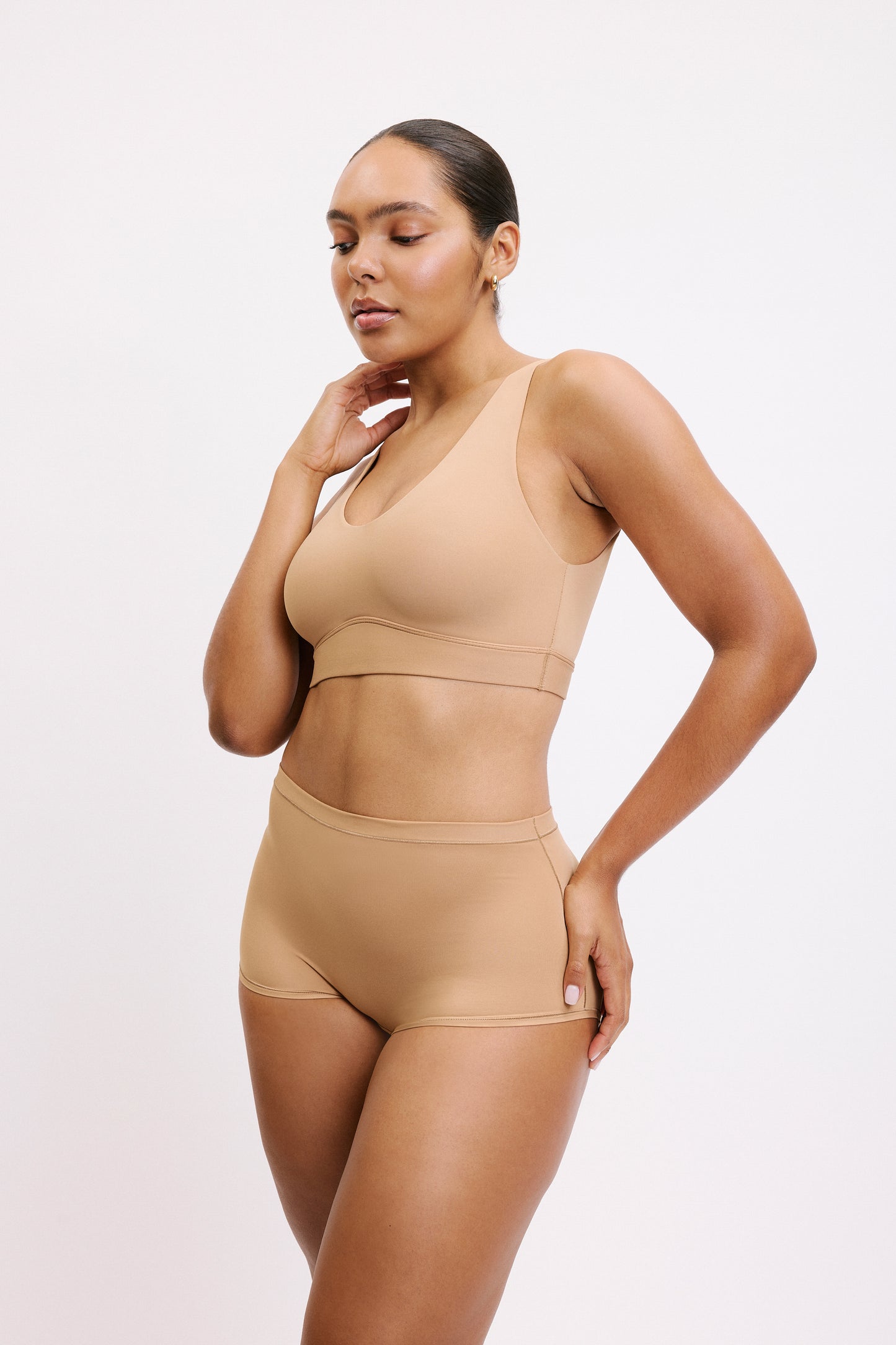 On a white studio backdrop, Amber is shown in a full-body three-quarter view wearing the Bliss Bralette in Tan with the matching brief, highlighting the bralette’s smooth underband and comfortable, everyday silhouette.