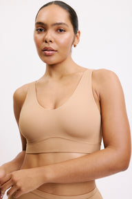 On a white studio backdrop, Amber is shown in a close-up front view wearing the Bliss Bralette in Tan, highlighting the scoop neckline, smooth cups, and soft microfiber fabric.