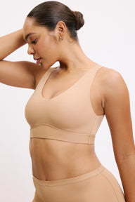 On a white studio backdrop, Amber is shown in a close-up side angle wearing the Bliss Bralette in Tan, highlighting the scoop neckline, supportive underband, and lightweight microfiber fabric. sizing: Amber wears size Medium